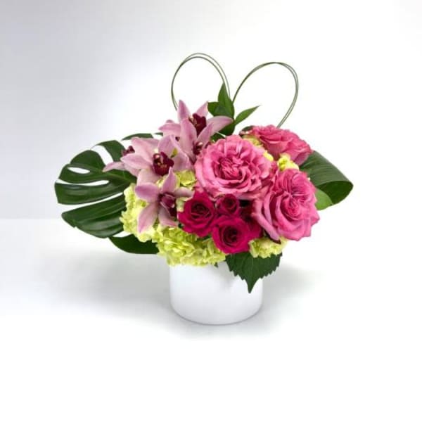 Low arrangement of pink roses, orchids, and green hydrangea in a white cylinder vase with a heart-shaped loop accent