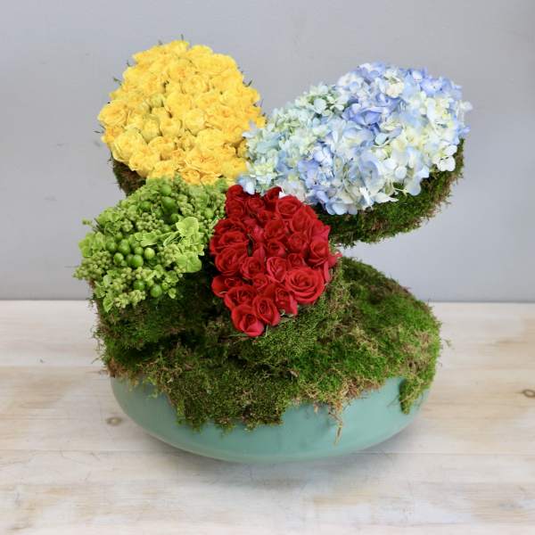 Modern moss-covered arrangement with yellow and red roses and blue hydrangeas in a low green bowl
