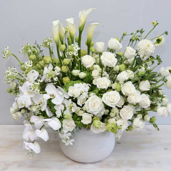 Large white and green arrangement of roses, calla lilies, orchids, and other blooms in a white ceramic bowl