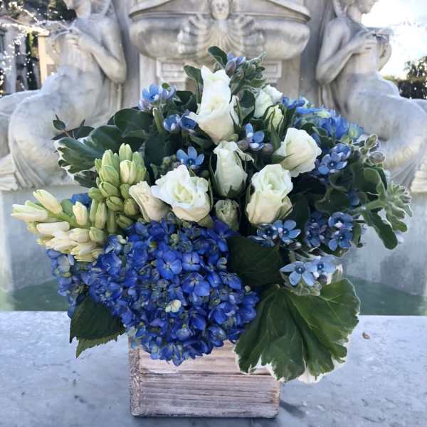 Compact blue and white flower arrangement with hydrangeas and roses in a rustic wooden cube vase.