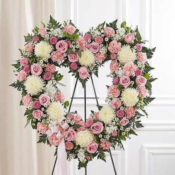 Heart-shaped standing wreath of pink roses, carnations, and white mums with a pink ribbon on an easel