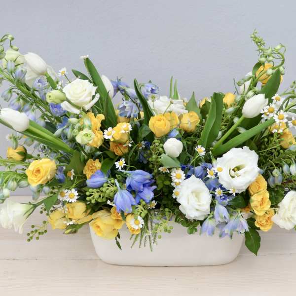 Low centerpiece of yellow roses, white tulips, and blue flowers in a white ceramic container