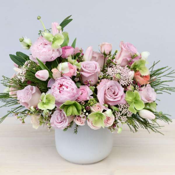 Low arrangement of soft pink roses, peonies, and tulips with light green blooms in a white ceramic pot