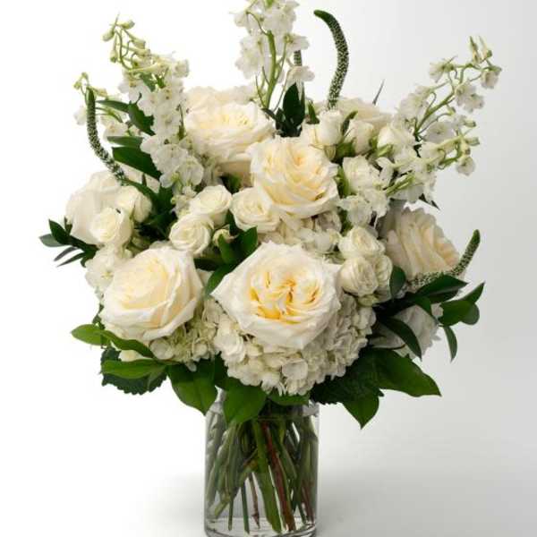 Tall arrangement of white and cream roses, hydrangeas, and delphinium in a clear glass vase