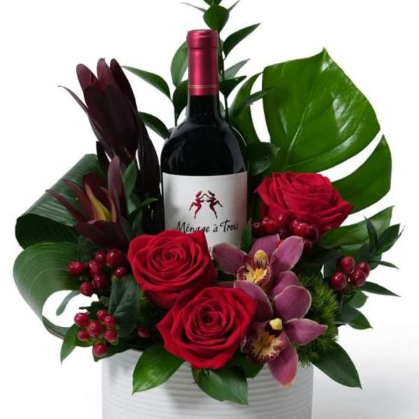 Red wine gift arrangement with red roses, pink orchids, berries, and large green leaves in a white round container
