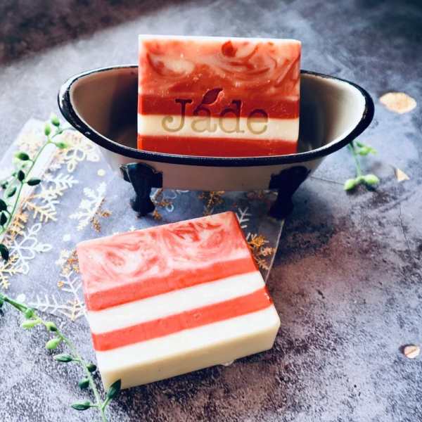 Striped red and white bar soaps, one in a small metal bathtub dish on a textured surface.