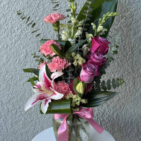 Tall pink and white arrangement with roses, lilies, and carnations in a glass vase with a pink ribbon.