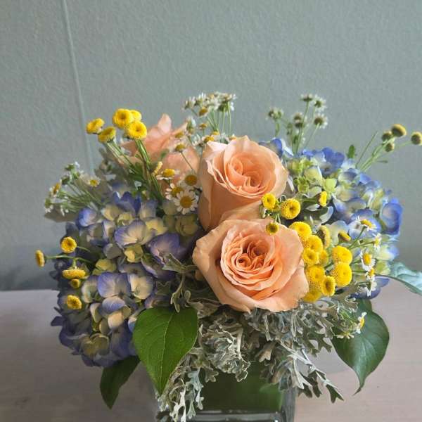 Low arrangement of peach roses, blue hydrangeas, and yellow and white daisies in a clear glass cube vase