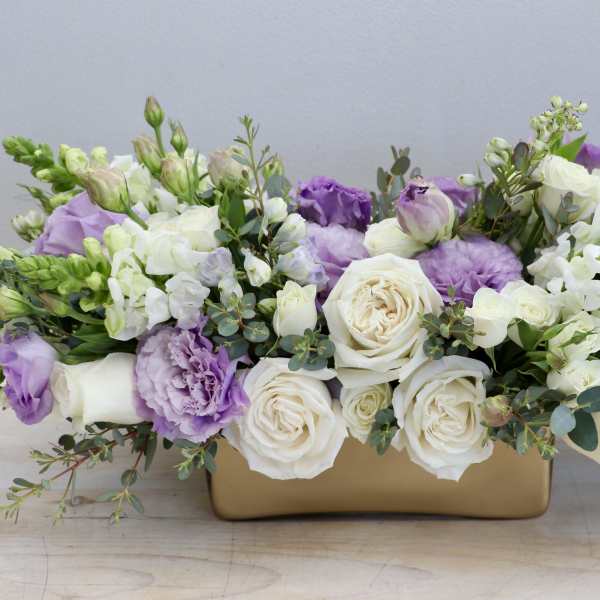 Low arrangement of white roses, purple lisianthus, and white snapdragons in a rectangular gold container.