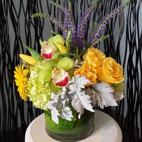 Compact yellow and green floral arrangement with orchids, roses, hydrangea and purple accents in a glass vase