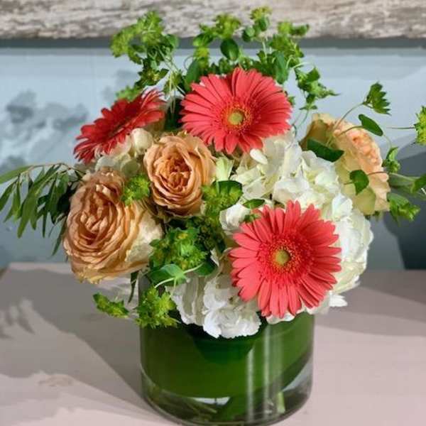 Short vase arrangement with coral gerbera daisies, peach roses, and white hydrangeas in a clear glass cylinder