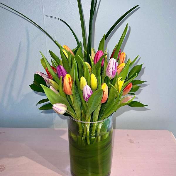 Mixed-color tulip arrangement with tall decorative grass in a clear glass cylinder vase.