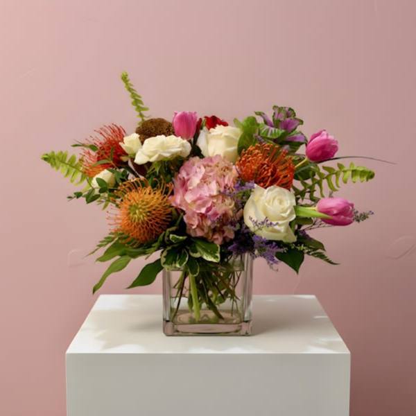 Mixed arrangement of pink tulips, white roses, hydrangea and orange protea in a clear glass cube vase