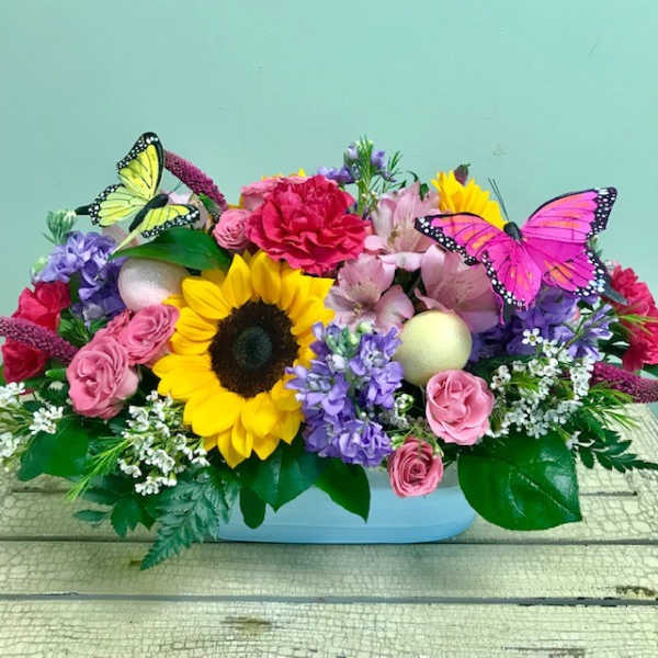 Low pastel centerpiece with sunflower, pink roses, purple stock and butterflies in a blue container