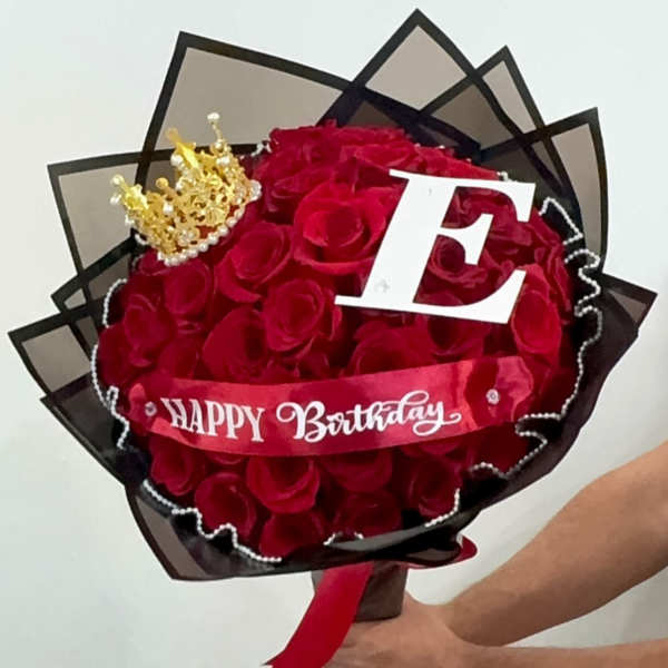 Bouquet of red roses wrapped in black paper with a crown, letter E, and Happy Birthday ribbon.