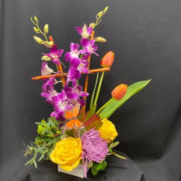 Tall arrangement with purple orchids, yellow roses, orange tulips, and a lavender mum in a white vase.