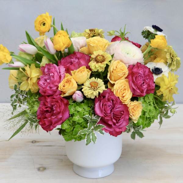 Bright arrangement of pink roses, yellow blooms, and green hydrangea in a white pedestal vase