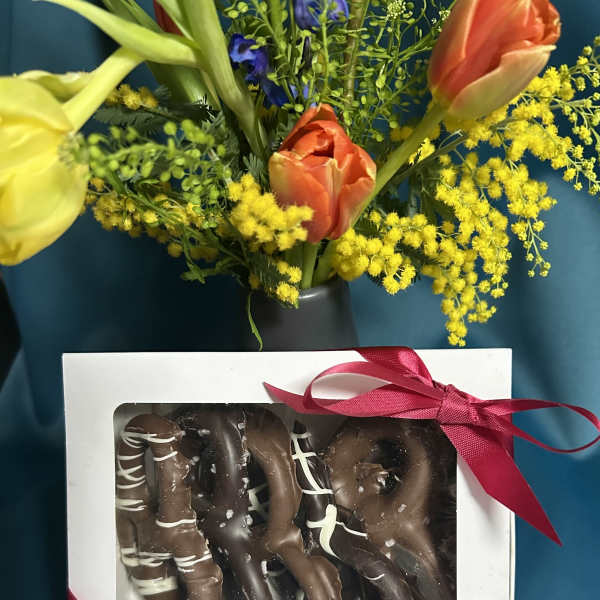 Gift set with yellow and orange tulips in a vase behind a box of chocolate-covered pretzels tied with a red ribbon.