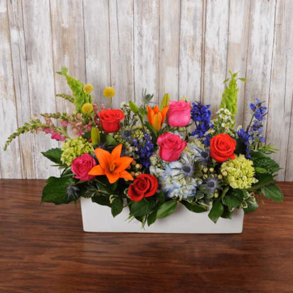 Rectangular centerpiece of bright mixed flowers including roses, lilies, hydrangeas, and delphinium in a white container