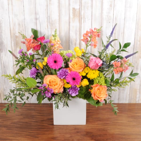 Bright mixed arrangement of pink gerbera daisies, orange roses, and yellow blooms in a white square vase