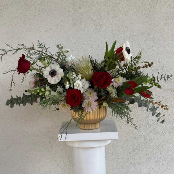 Asymmetrical arrangement of red roses and white flowers in a gold compote vase on a white pedestal.
