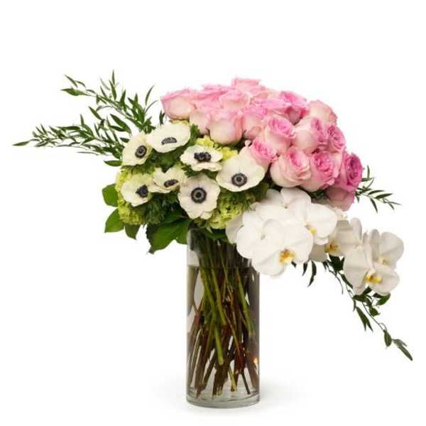 Asymmetrical arrangement of pink roses, white anemones, and orchids in a tall clear glass cylinder vase.