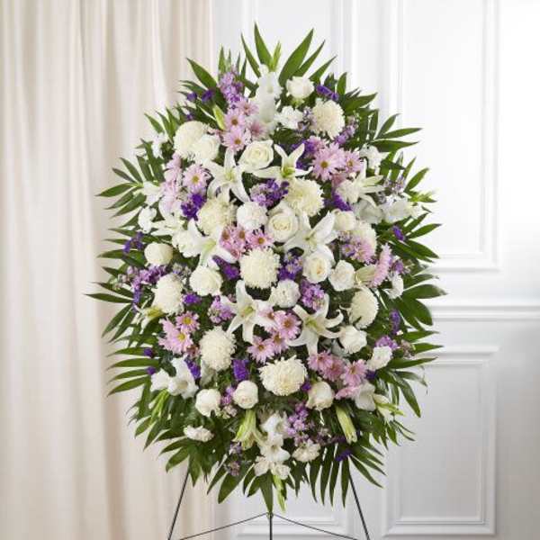 Tall standing spray of white lilies and roses with pink and purple flowers framed by green leaves on an easel.