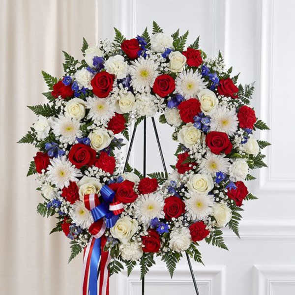 Standing wreath of red and white roses and mums with blue accents and a patriotic ribbon bow.