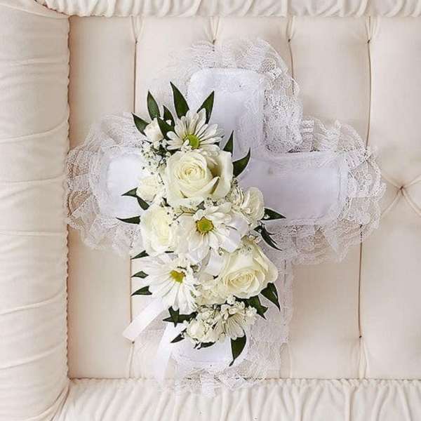 White floral cross pillow with roses and daisies resting on a cream tufted casket interior.