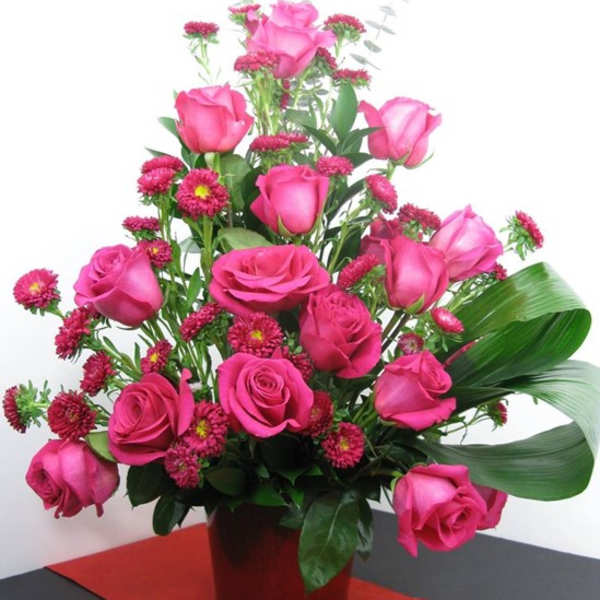 Tall arrangement of hot pink roses and small pink asters in a red pot on a red table runner