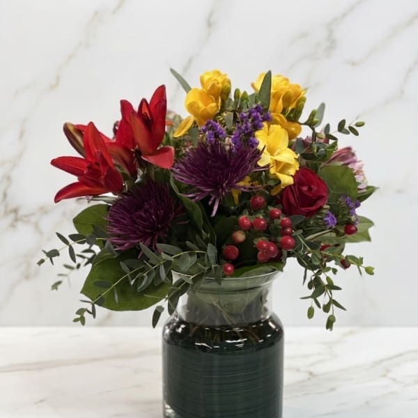 Bright mixed bouquet with red lilies, yellow flowers, and purple blooms in a green glass vase