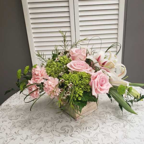 Low centerpiece of pink roses, pink carnations, green hydrangea and white lilies in a wooden cube vase
