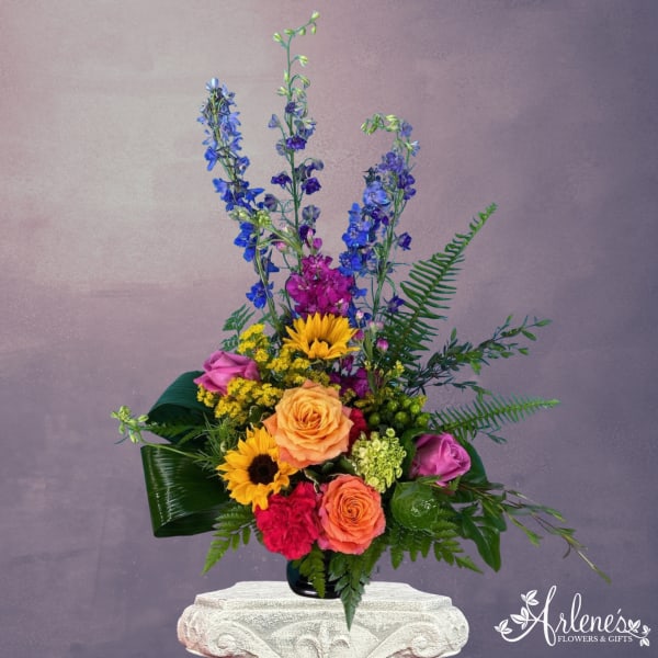 Tall mixed arrangement with blue delphinium, yellow sunflowers, orange roses, and pink blooms in a black vase.