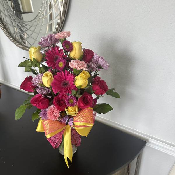 Bright mixed bouquet of pink roses, gerbera daisies, and yellow roses in a clear vase with a yellow and pink bow.