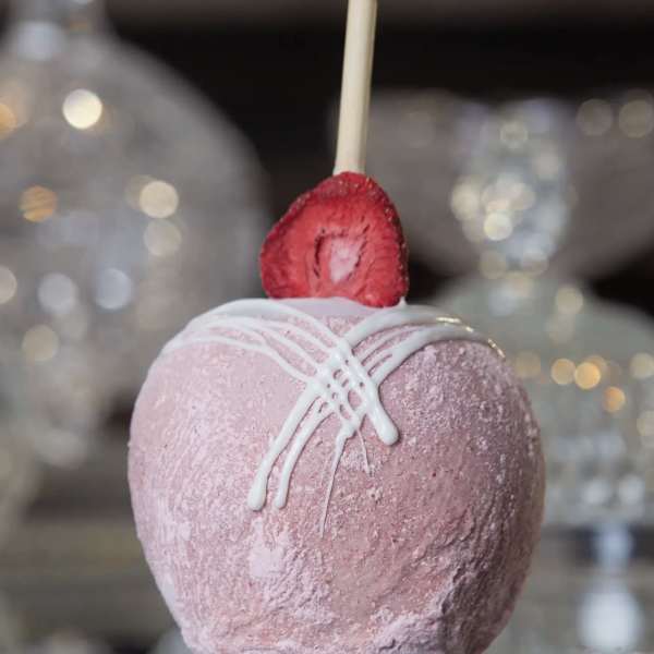Pink candy-coated apple with white drizzle and a strawberry slice on top, set on a reflective surface.