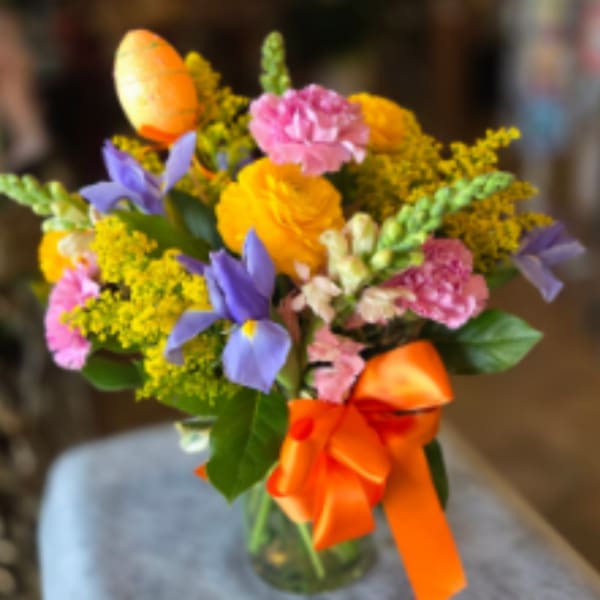 Bright mixed bouquet of yellow, pink, and purple flowers in a clear vase with an orange bow and egg pick.