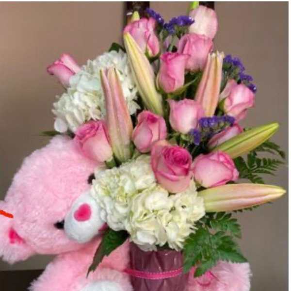 Pink teddy bear hugging a vase of pink roses, lilies, and white hydrangeas