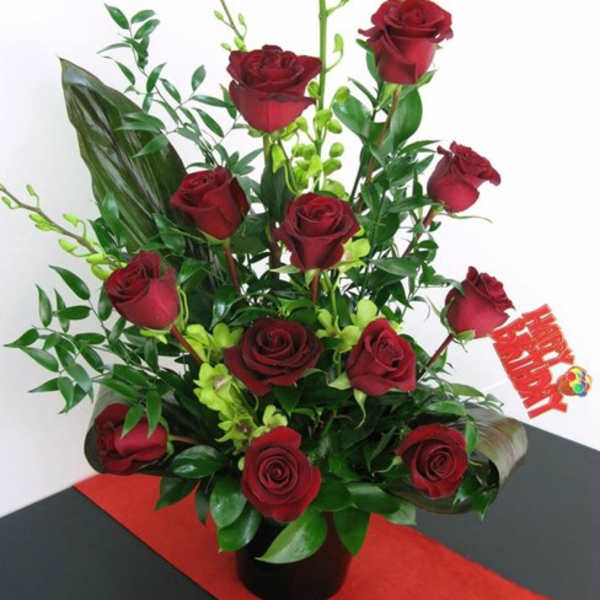 Tall arrangement of dark red roses with green orchids in a black vase and a red Happy Birthday pick