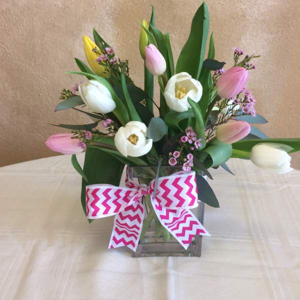 Arrangement of pink, white, and yellow tulips with small pink blooms in a glass cube vase with a chevron bow