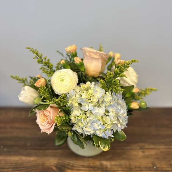 Low arrangement of blue hydrangea, white ranunculus, and peach roses in a round white vase.