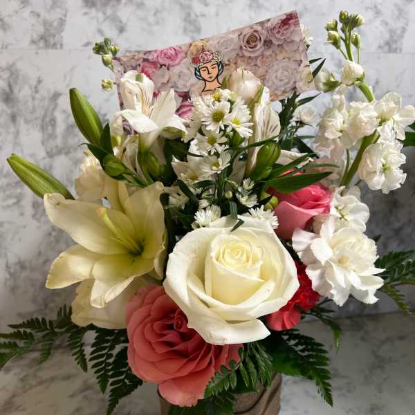 Mixed bouquet of white lilies, roses, and pink blooms in a burlap-wrapped cube vase with a floral card.