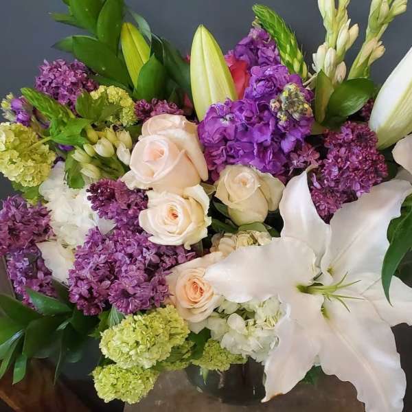Mixed arrangement of purple lilacs, white lilies, and pale roses in a clear glass vase