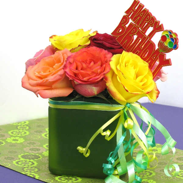 Bright mixed roses in a green cube vase with a Happy Birthday sign and curling ribbons.