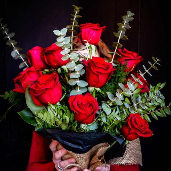 Hand-tied bouquet of red roses wrapped in burlap and black paper, held against a dark background