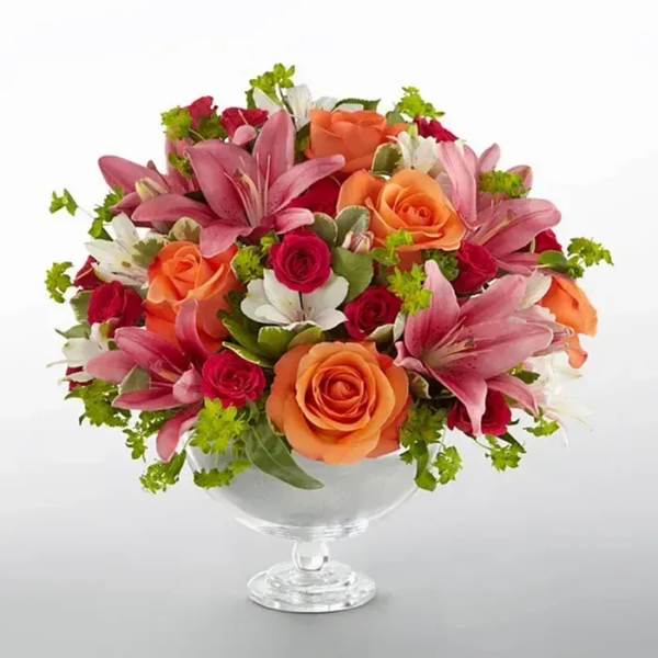 Low arrangement of pink lilies with orange and red roses in a clear glass pedestal bowl