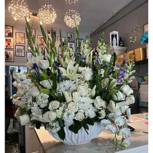 Large white floral arrangement with lilies, roses, and orchids in a white bowl with touches of blue flowers.