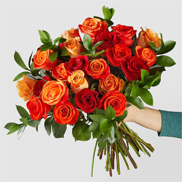 Handheld bouquet of orange, red, and peach roses with lush green accents against a light background