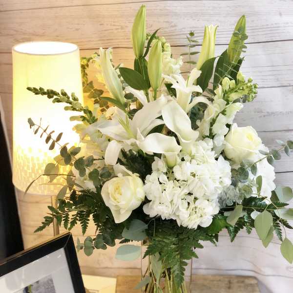 Tall white lily, rose, and hydrangea arrangement with greenery in a clear glass vase on a wooden table