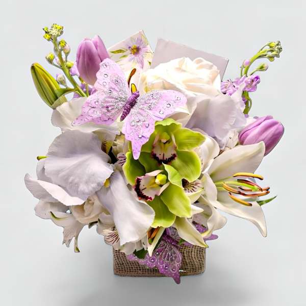 Square vase arrangement of white lilies, green orchids, roses, and lavender tulips with pink butterfly accents