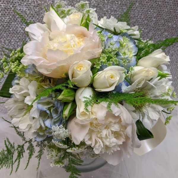 Round bouquet of ivory peonies, white roses, and pale blue flowers in a glass vase.